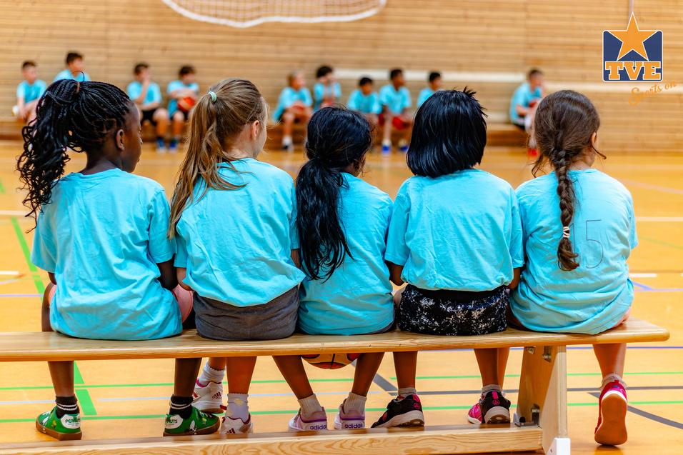 Fünf Kinder in blauen T-Shirts sitzen auf einer Bank und schauen auf eine Gruppe von Spielern in einem Sportraum.