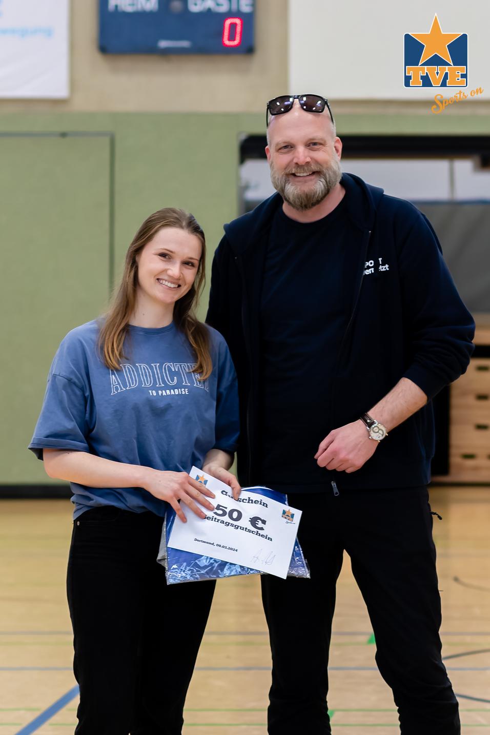 Frau und Mann halten ein Preisschild mit „50 €“ in einer Sporthalle, im Hintergrund die Anzeige des Spiels.