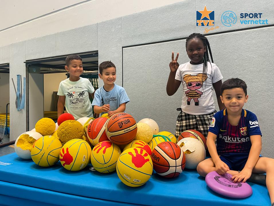 Integration SV Vier Kinder stehen um einen Haufen von verschiedenen Bällen auf einer blauen Turnmatte in einer Sporthalle.