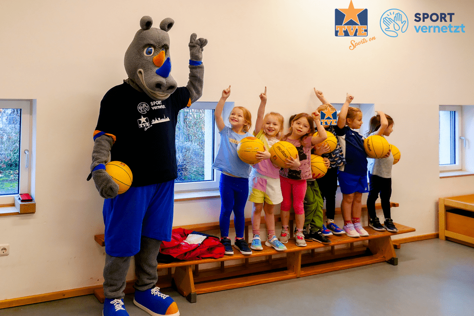 TVE x Kita Eine lustige Maskottchenfigur steht neben mehreren Kindern, die Basketballbälle halten und fröhlich in die Kamera zeigen.
