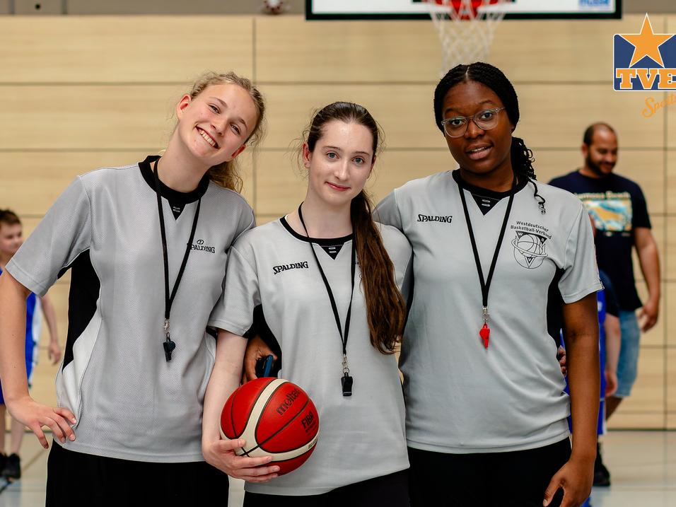 Drei Basketballtrainerinnen in grauen Trikots stehen lächelnd mit einem Basketball in der Hand auf dem Spielfeld.