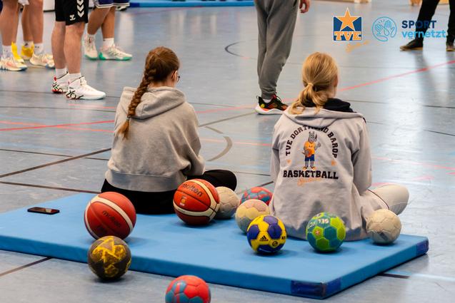 Zwei Jugendliche sitzen auf einem blauen Mattenst&uuml;ck, umgeben von verschiedenen Sportb&auml;llen in einer Sporthalle.