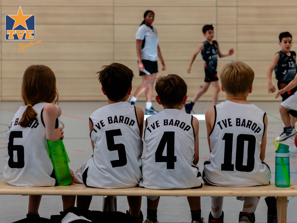 Vier Kinder in Basketballtrikots sitzen auf einer Bank und beobachten ein Spiel in einer Sporthalle.