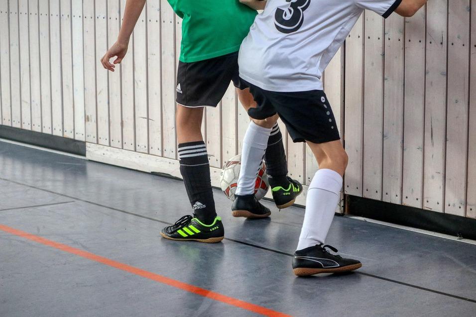 Zwei Fußballspieler in Sportkleidung dribbeln den Ball an einer Wand in einer Halle entlang.