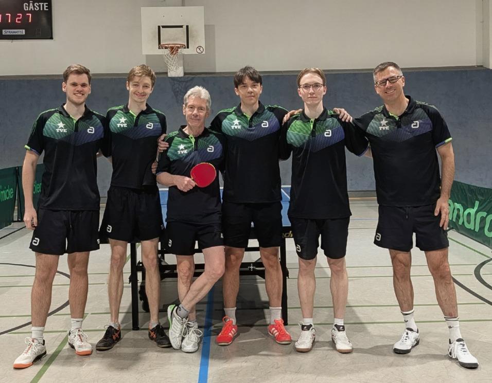 Sechs Tischtennisspieler in Teamkleidung posieren lächelnd vor einem Tischtennistisch in der Sporthalle.
