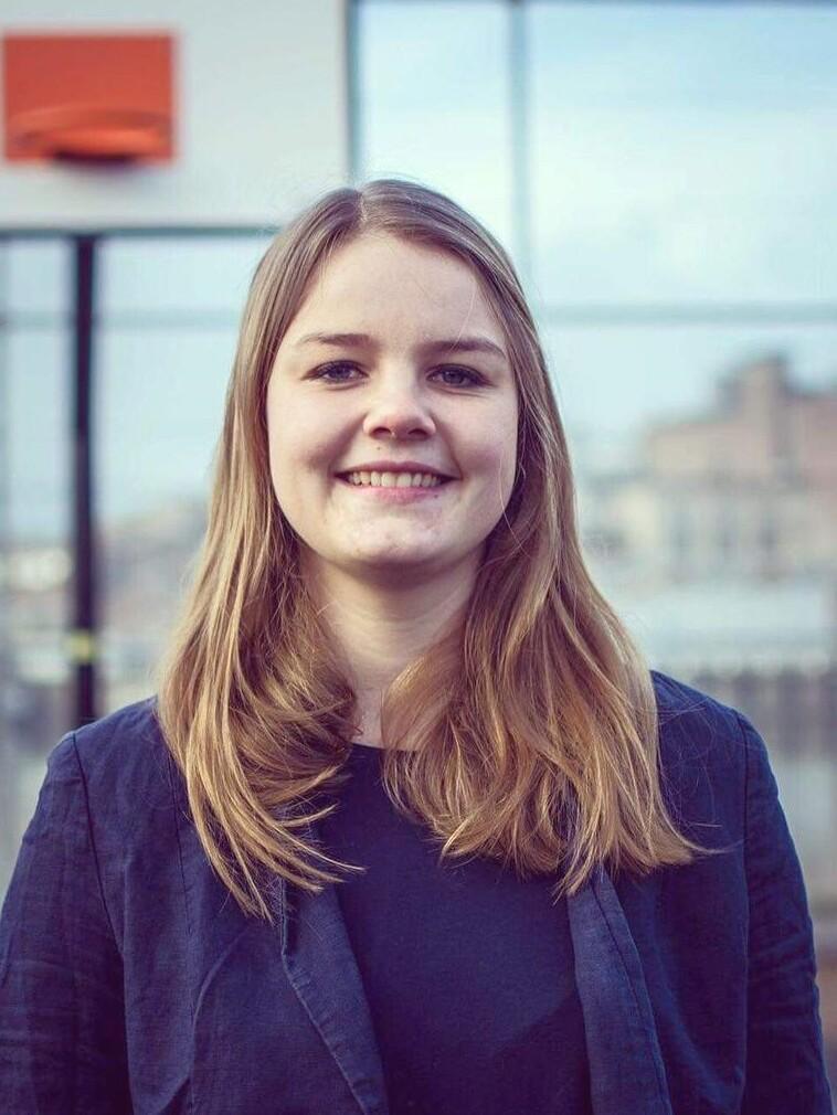 Lächelnde Frau mit langen, blonden Haaren, im Vordergrund eines großen Fensters, hinter ihr ist ein Basketballkorb sichtbar.