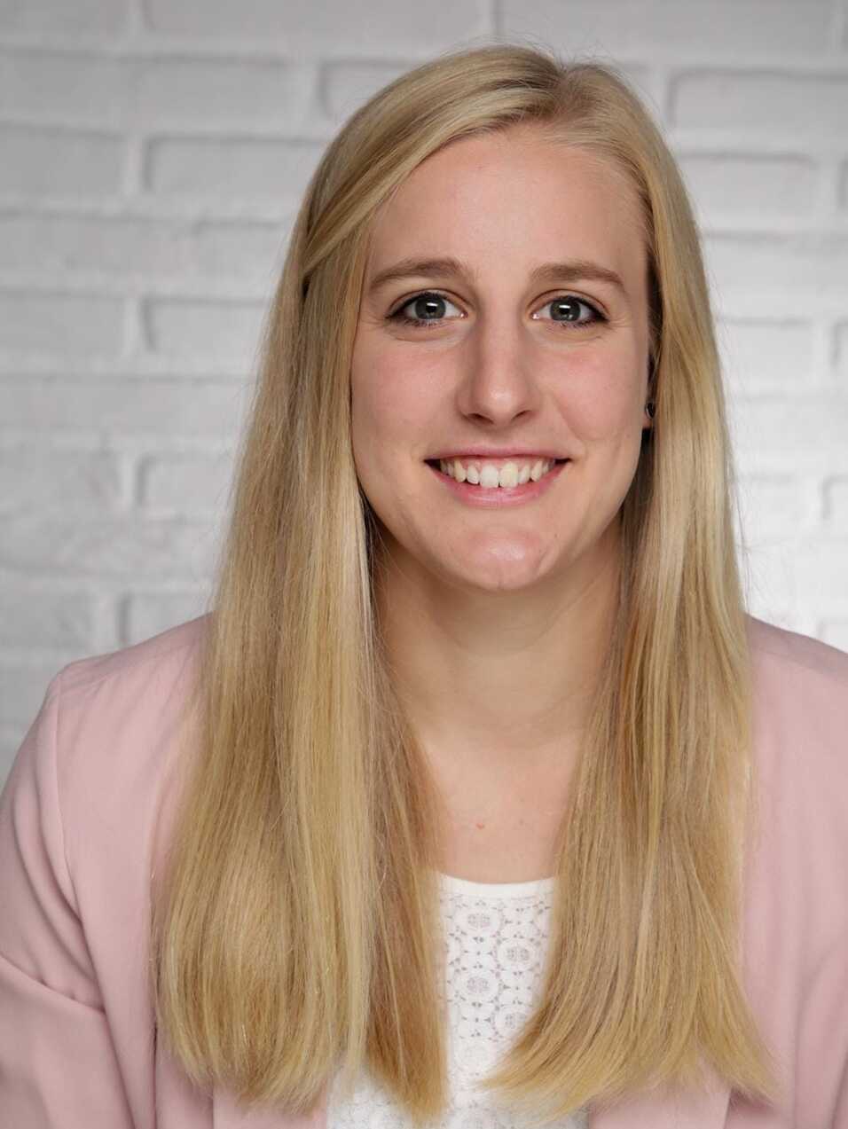 Porträt einer lächelnden Frau mit blonden Haaren in einer hellen Jacke vor einer weißen Backsteinwand.