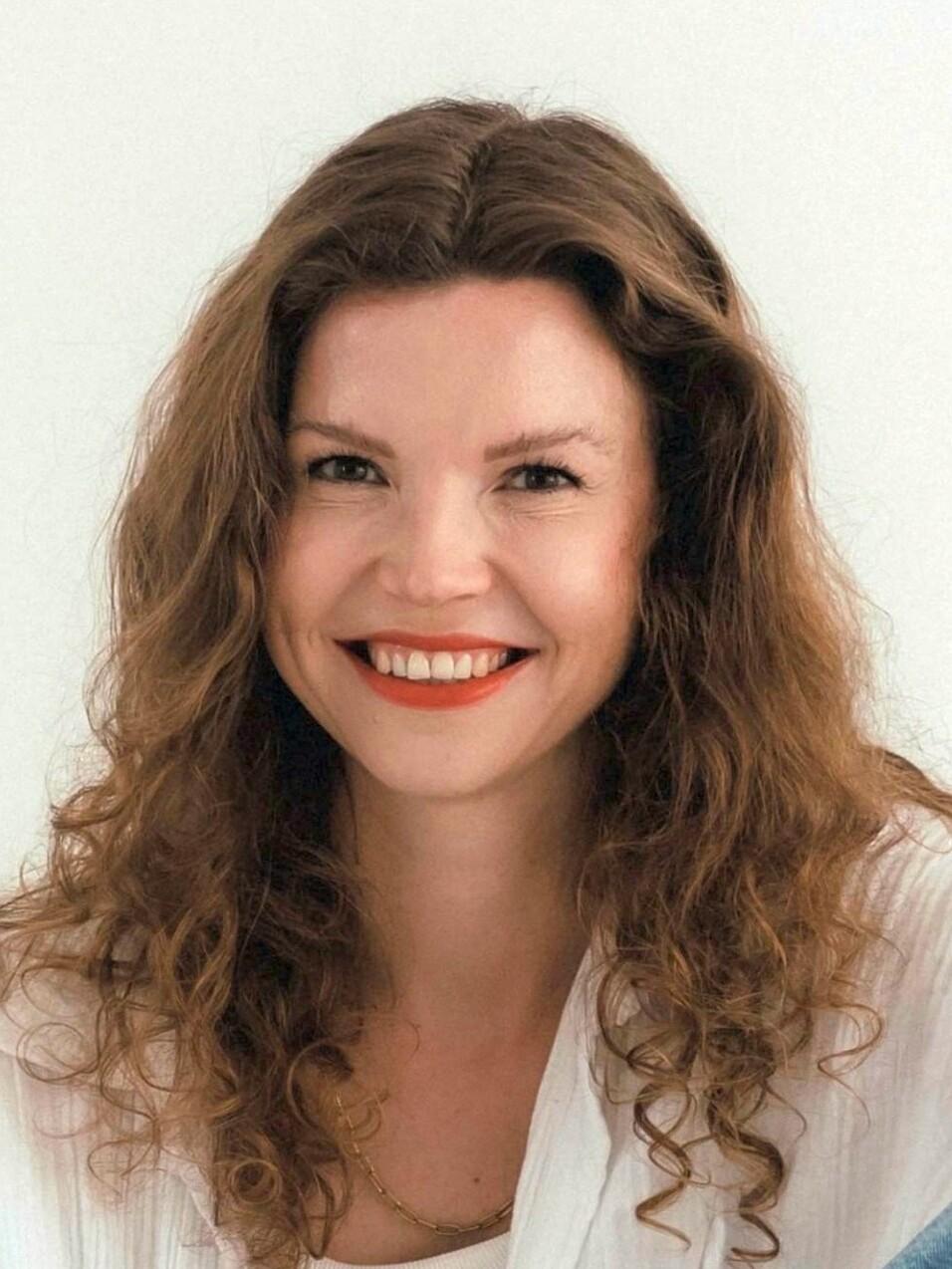 Lächelnde Frau mit lockigem, braunem Haar und rotem Lippenstift vor neutralem Hintergrund.