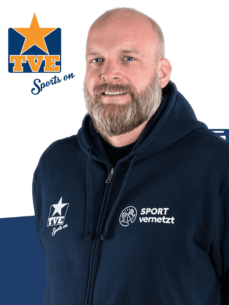 Nahaufnahme eines lächelnden Mannes mit Vollbart in blauer Jacke mit Logos von TVE und SPORT vernetzt.