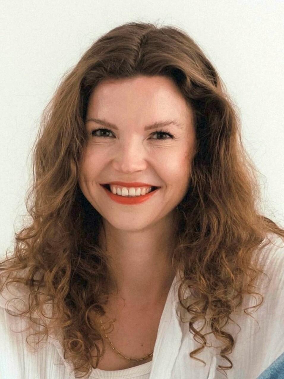 Lächelnde Frau mit lockigem, braunem Haar und rotem Lippenstift vor neutralem Hintergrund.