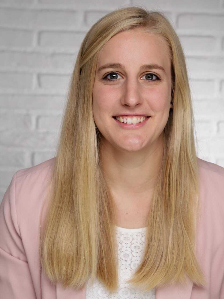 Porträt einer lächelnden Frau mit blonden Haaren in einer hellen Jacke vor einer weißen Backsteinwand.