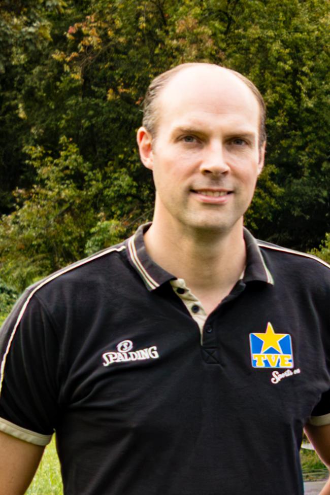 Mann mit kurzem Haar trägt ein schwarzes Polohemd mit Aufdruck und steht vor einer grünen, bewaldeten Kulisse.