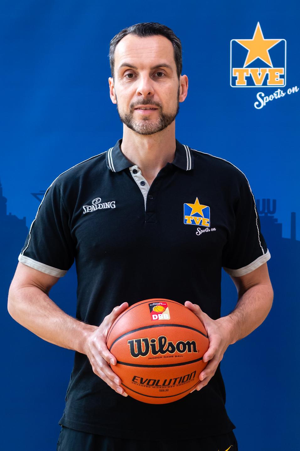 Porträt eines Mannes mit Basketball in der Hand, trägt schwarzes Polo mit Logo auf blauem Hintergrund.