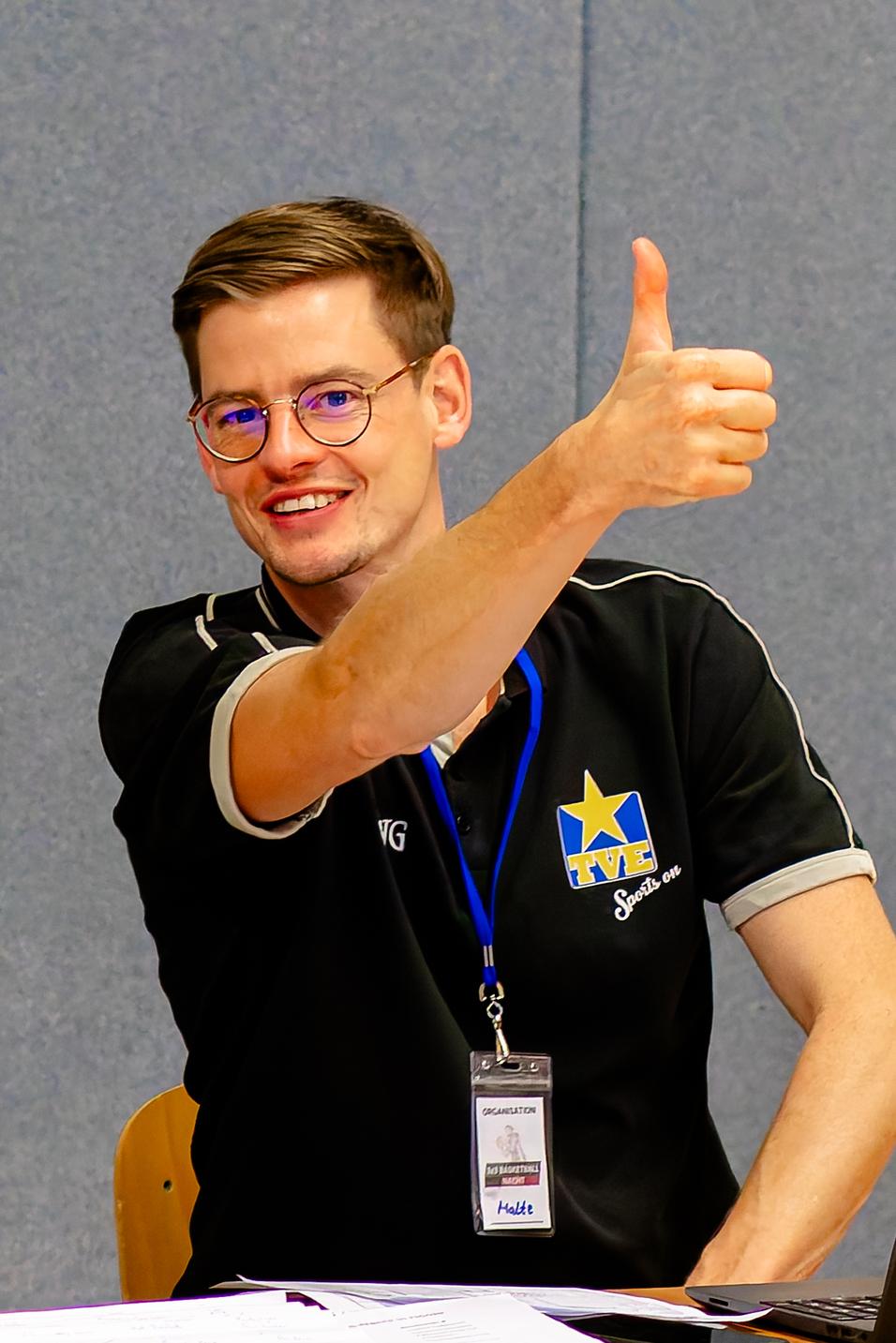 Lächelnder Mann in schwarzem Shirt zeigt Daumen hoch, sitzt vor Tisch mit Zetteln und Ausweis.
