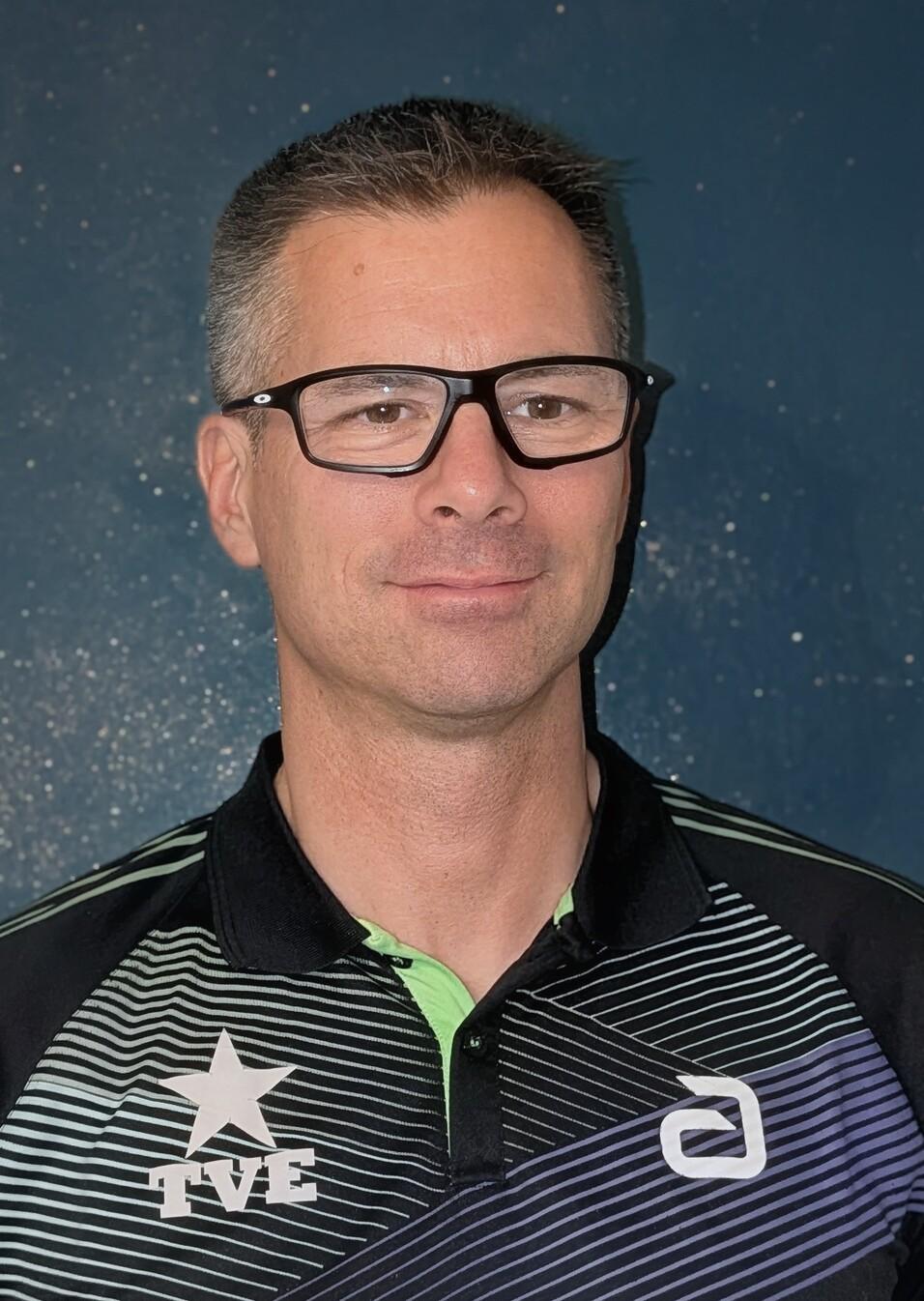 Porträt eines Mannes mit Brille, der ein schwarz-grünes Sportoberteil trägt, vor einem blauen Hintergrund.