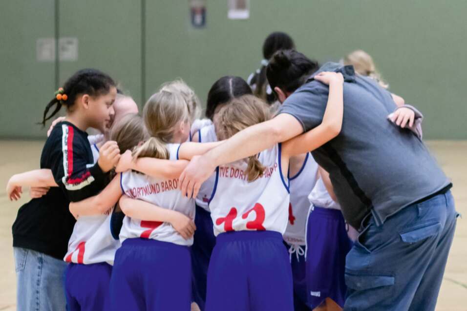 M&auml;dchen-Basketballmannschaft umarmt sich vor einem Spiel, Trainer steht in der Mitte, alle tragen einheitliche Trikots.