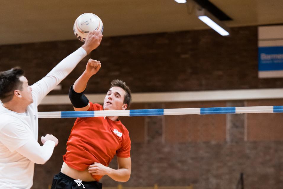Zwei Spieler im Hallenvolleyball springen, um einen Ball über das Netz zu schlagen, während andere im Hintergrund trainieren.